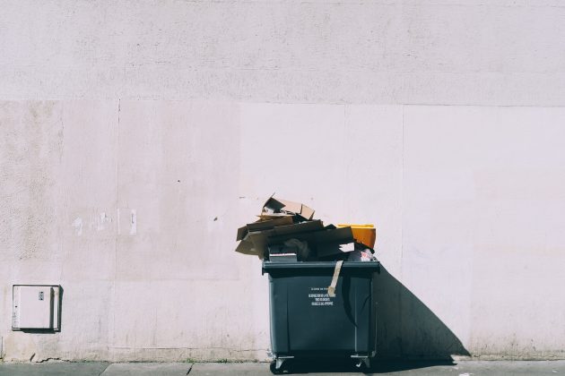 Dark green dumpster filled with cardboard boxes against white wall