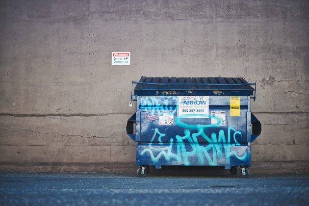 Blue front-load dumpster with graffiti parked near concrete wall
