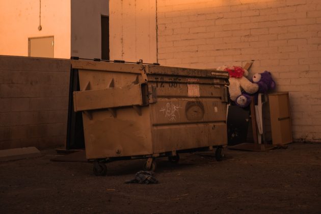Beige roll-off dumpster and discarded teddy bears in alleyway